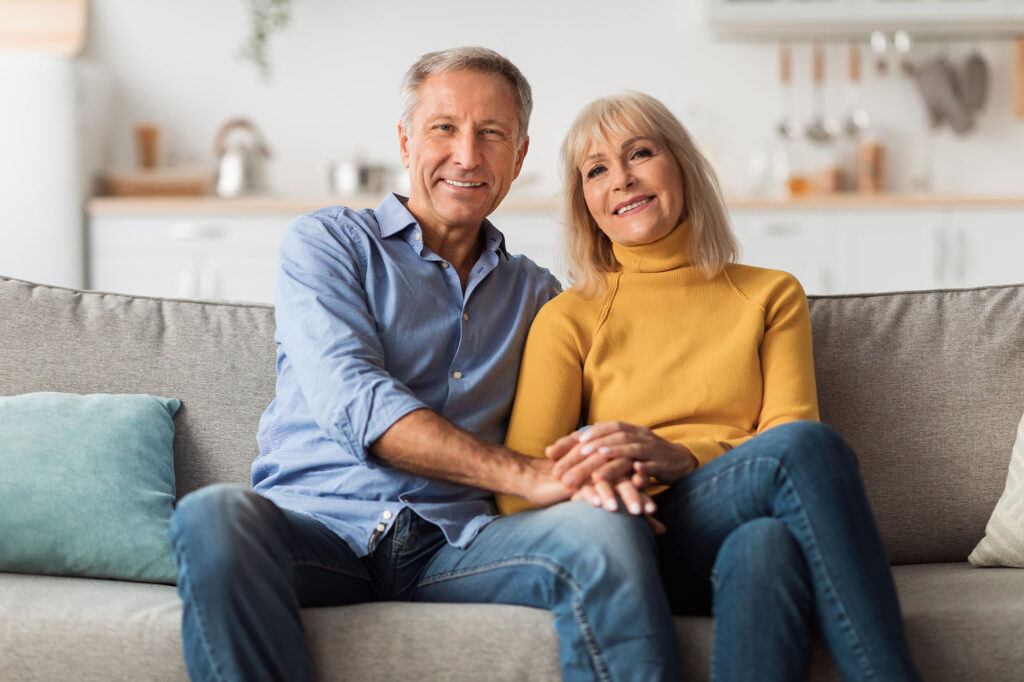 Older Spouses Holding Hands Sitting On Sofa Posing At Home